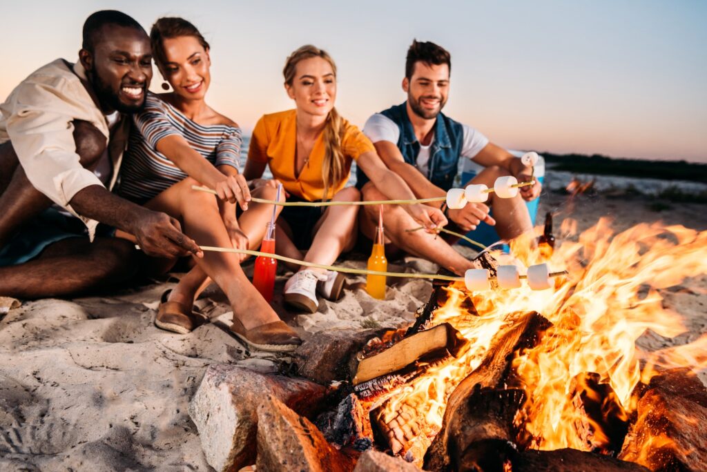 happy young multiethnic friends roasting marshmallows at bonfire on beach